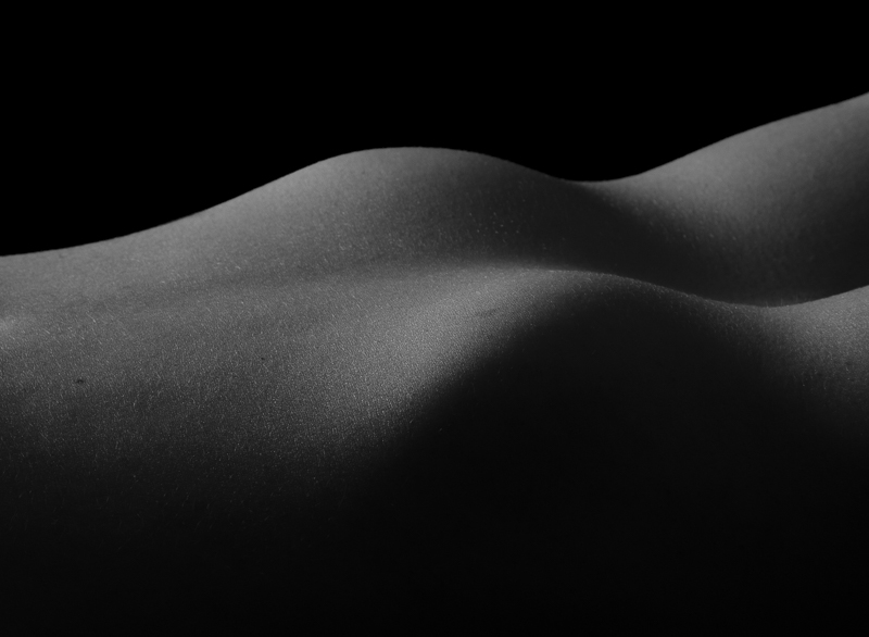 This black and white abstract view of the human form resembles undulating sand dunes. The image is softly lit from the left and the structure of the dunes fade into shadow.